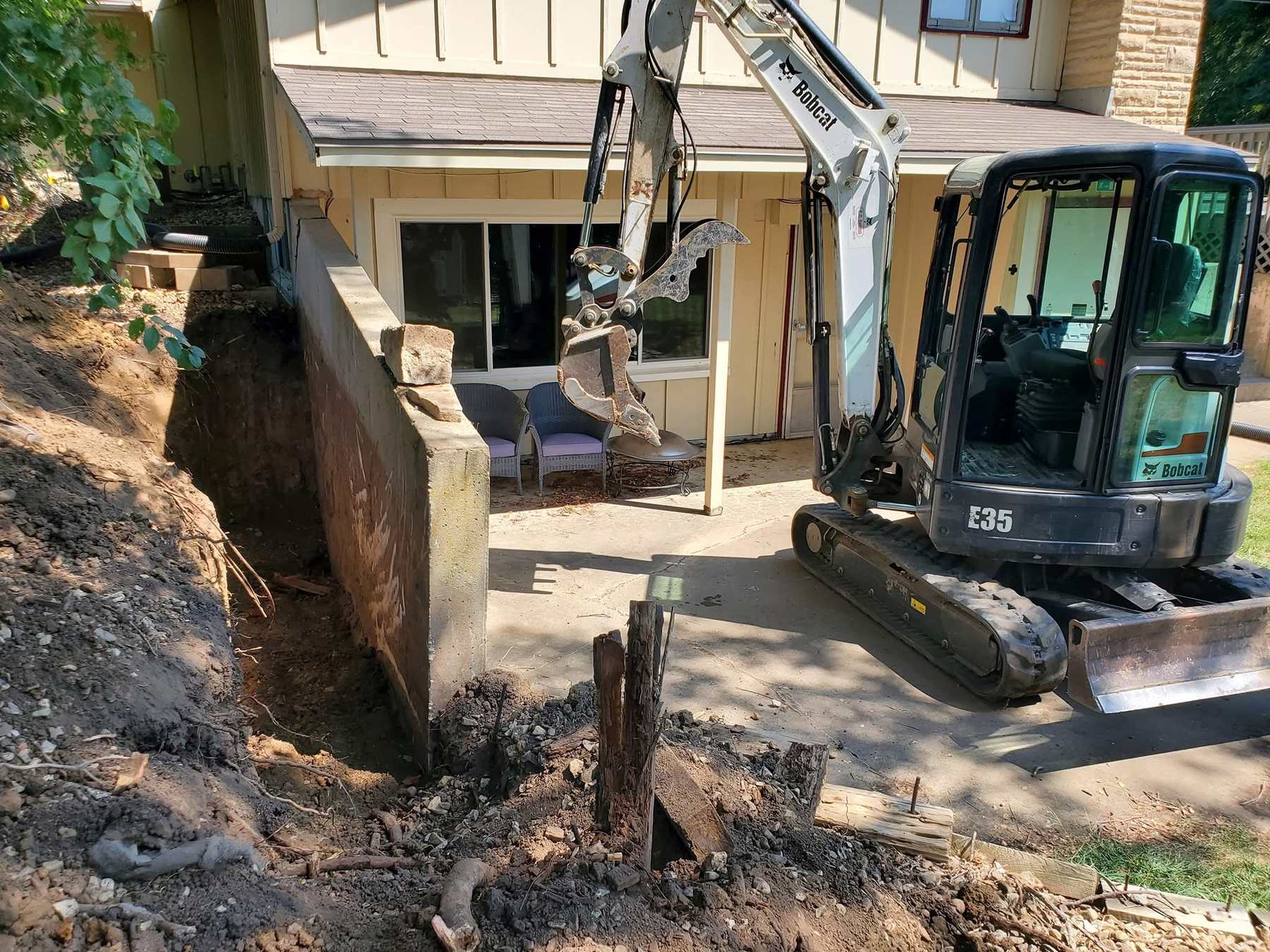 Bobcat E35 mini excavator with a grapple digging beside a concrete retaining wall on a Kansas foundation job